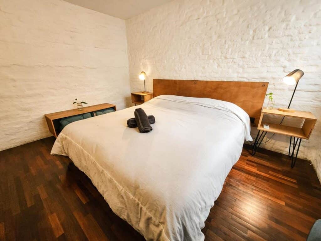 Quarto do MedioMundo Hostel. Uma cama de casal no centro, de cada lado uma estante de apoio com um abajur, no fundo uma pequena cômoda. Imagem para ilustrar post sobre hostels em Montevidéu.