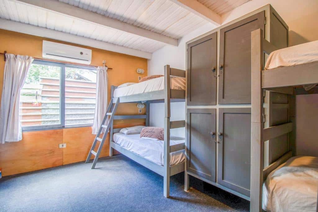 A imagem mostra um quarto com duas beliches encostadas na parede direita. Entre as camas, há um grande guarda-roupa cinza com portas fechadas. O quarto tem uma parede e teto bege, com acabamento branco no topo da parede onde ela encontra o teto. O chão é coberto por carpete azul. Há uma janela na equerda com cortinas parcialmente fechadas, permitindo a entrada de luz natural. Também há um aparelho de ar-condicionado instalado na parede próxima a janela. Está representando o post sobre hostels em Punta Del Este.