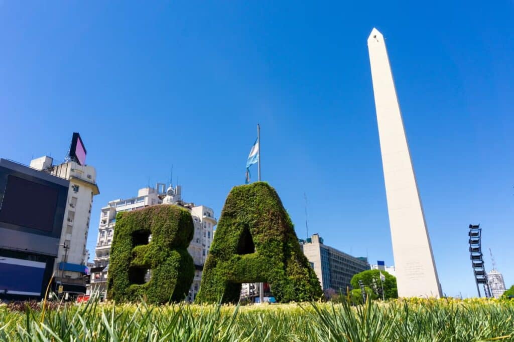 A imagem mostra o icônico Obelisco de Buenos Aires, um dos marcos mais famosos da capital argentina. O obelisco é uma estrutura alta e esguia de cor branca, destacando-se contra um céu azul claro. À frente do obelisco, há letras grandes cobertas de vegetação que formam a sigla "BA", representando "Buenos Aires". Uma bandeira da Argentina tremula no mastro atrás das letras. Ao redor, é possível ver edifícios e construções típicas da área urbana central da cidade. A imagem foi capturada em um dia ensolarado, realçando as cores vivas do local.