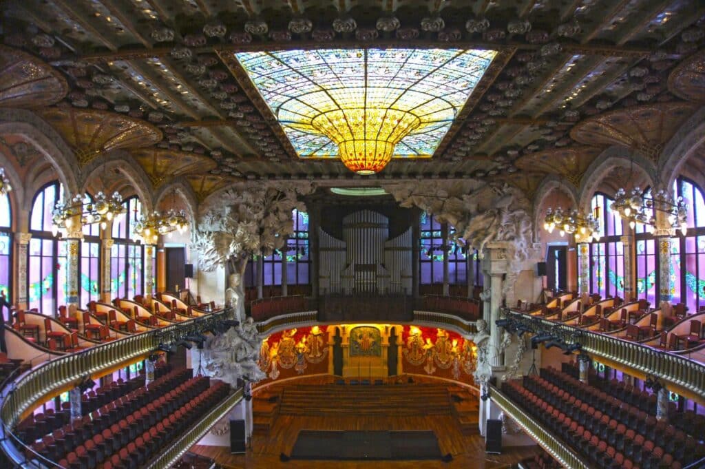 A imagem mostra o interior do Palácio da Música Catalã, uma das opções de o que fazer em Barcelona, com destaque para seu teto ornamentado com vitrais coloridos formando uma cúpula central em formato de flor. As paredes laterais possuem grandes janelas em arcos, também decoradas com vitrais. No centro do palco, um órgão impressionante se impõe, enquanto as laterais estão adornadas com esculturas e detalhes intrincados. As cadeiras do auditório estão dispostas em fileiras, divididas em dois níveis com varandas curvadas, complementando o ambiente de estilo modernista.