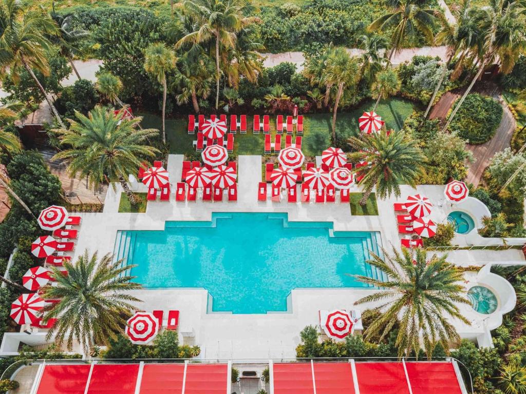 Vista aérea da piscina do Faena Hotel Miami Beach cercada por guarda-sóis vermelhos e brancos, espreguiçadeiras vermelhas, palmeiras e vegetação paisagística.