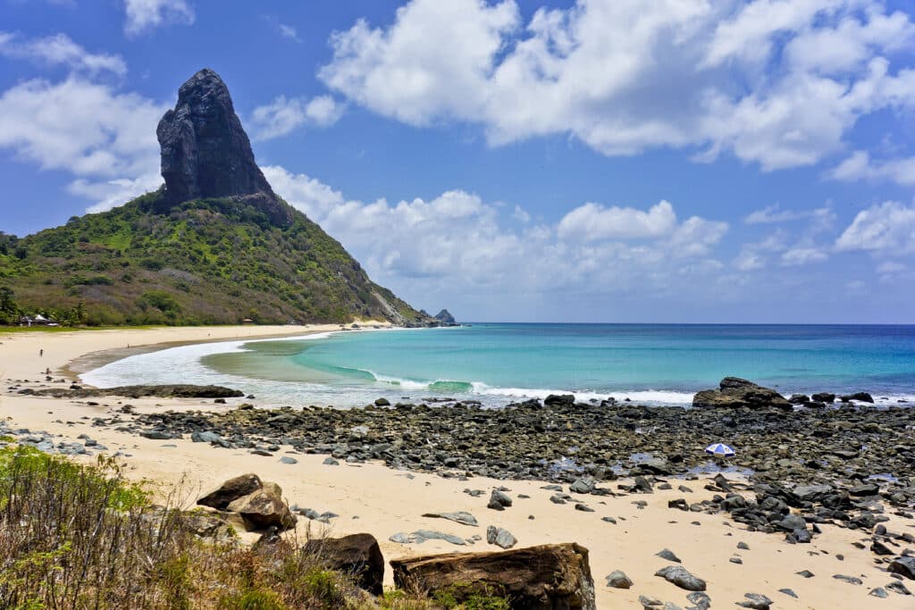 Praia da Conceição que ilustra o post de o que fazer em Fernando de Noronha. A imagem mostra uma paisagem de praia cênica com uma colina ou montanha íngreme ao fundo. O céu está principalmente claro, com algumas nuvens dispersas. O oceano parece calmo, com diferentes tons de azul e turquesa. No primeiro plano, há uma praia de areia intercalada com áreas rochosas e alguma vegetação. Um único guarda-sol azul é visível na areia, sugerindo um local tranquilo e possivelmente isolado.