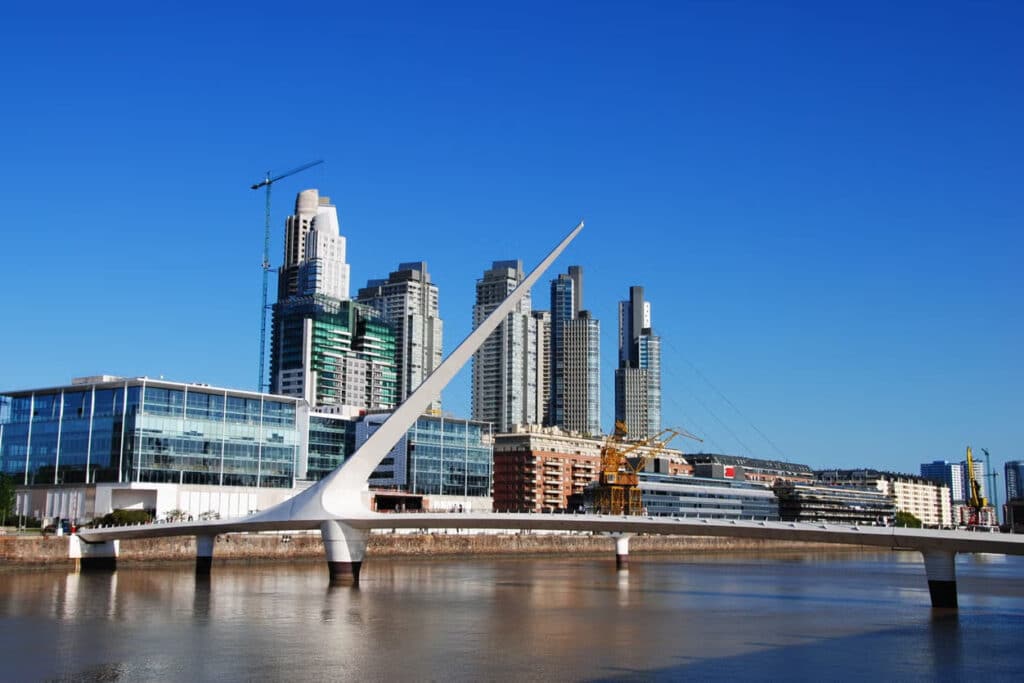 A imagem mostra a famosa Ponte da Mulher (Puente de la Mujer), localizada em Puerto Madero, Buenos Aires. Esta ponte pedonal, projetada pelo arquiteto espanhol Santiago Calatrava, é conhecida por seu design moderno e elegante, com uma estrutura inclinada que lembra a forma de uma vela ou uma dança. Ao fundo, é possível ver edifícios altos e modernos que compõem o horizonte de Puerto Madero, um dos bairros mais novos e sofisticados da cidade. A ponte atravessa o canal e se destaca pelo contraste entre sua cor branca e o azul intenso do céu.