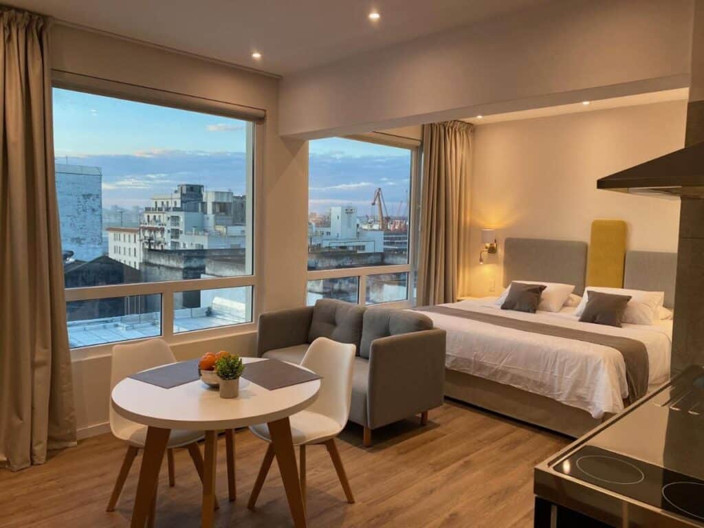 Flat do Puertovideo. Uma cama de casal no fundo, na frente um sofá e uma mesa de jantar. No canto do lado esquerdo as paredes de vidro e do lado direto uma minicozinha.