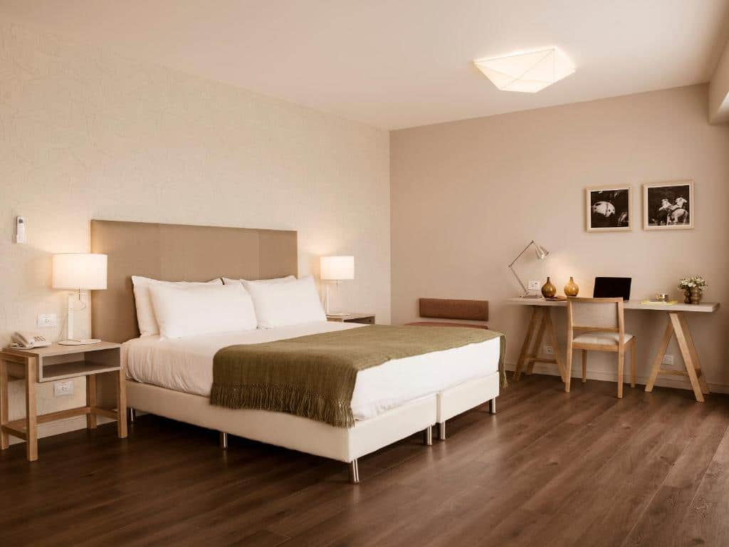 O quarto do hotel Dazzler by Wyndham Polo, em Buenos Aires, apresenta um design moderno e aconchegante. A cama de casal é grande e confortável, com uma cabeceira estofada em tom bege claro. A roupa de cama é branca, proporcionando um visual limpo e elegante, complementado por uma manta verde musgo aos pés da cama.
Em ambos os lados da cama, há mesas de cabeceira brancas com luminárias modernas, oferecendo uma iluminação suave e agradável para o ambiente. À direita da cama, um espaço de trabalho funcional é composto por uma mesa de madeira clara e uma cadeira combinando. O espaço é decorado com duas fotografias em preto e branco na parede, adicionando um toque artístico ao ambiente.
O piso é de madeira em tom escuro, que contrasta de forma elegante com as paredes claras e os móveis em cores neutras. A iluminação do quarto é complementada por uma luminária de teto moderna, que proporciona uma luz suave e difusa.