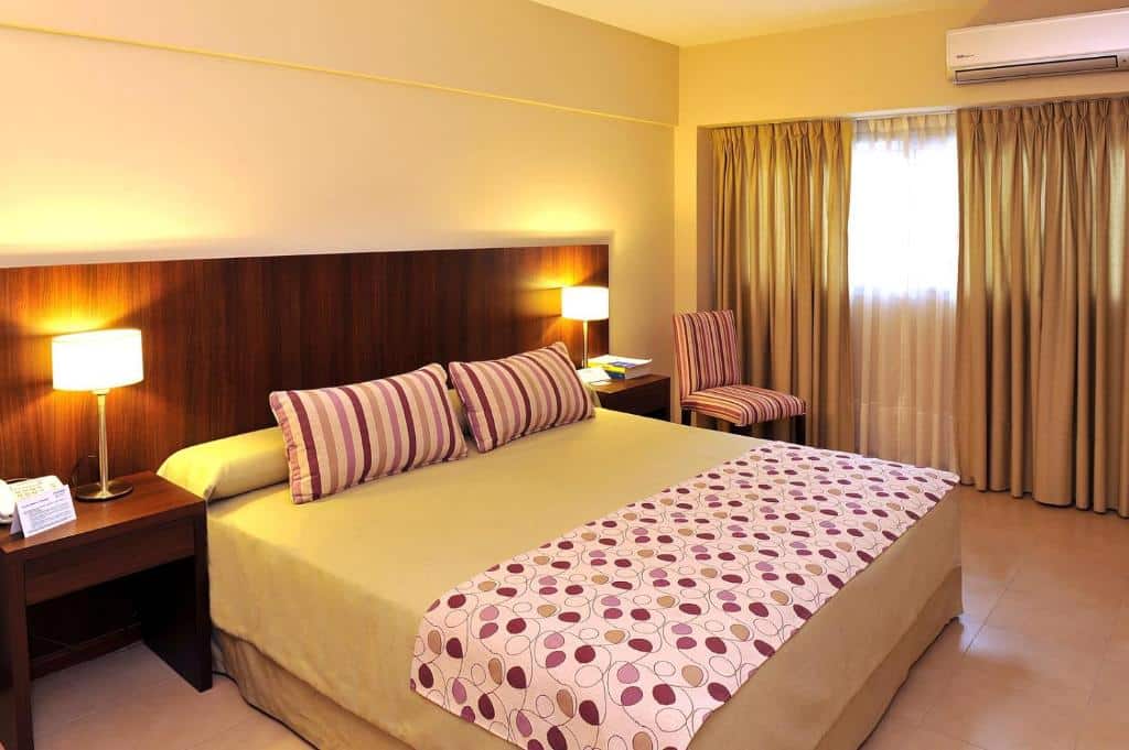 A imagem mostra um quarto do Hotel Intersur San Telmo, decorado de forma simples e aconchegante. A cama é grande, com roupa de cama em tons neutros e uma manta decorativa com estampa geométrica. Há dois abajures sobre mesas de cabeceira de madeira escura, que combinam com o painel da cabeceira da cama. O quarto tem uma cadeira estofada listrada ao lado de uma janela com cortinas longas, que permitem a entrada de luz natural. O ambiente transmite uma sensação de conforto e praticidade, ideal para descanso.