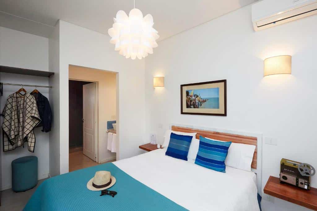 A imagem mostra um quarto do L'Adresse Hôtel Boutique decorado de maneira elegante e acolhedora. O quarto tem uma cama de casal com lençóis brancos e uma colcha azul clara. Acima da cama, há um quadro com uma imagem de uma praia. As paredes são brancas, criando uma sensação de amplitude e luminosidade. Ao lado da cama, há mesas de cabeceira de madeira com luminárias de parede modernas. Uma luminária de teto com design floral pendente completa a iluminação suave do ambiente. O quarto também possui um armário aberto com cabides e um pequeno pufe azul.