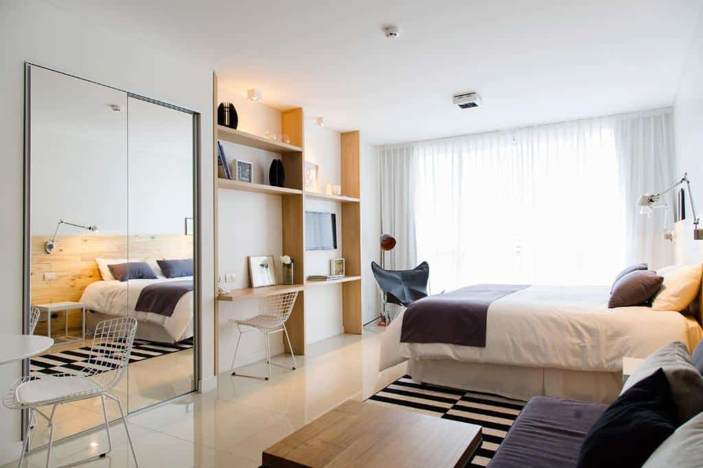 A imagem mostra um quarto moderno e bem iluminado do Lemon Suites. O espaço é amplo, com uma cama de casal centralizada, coberta com roupas de cama brancas e um cobertor cinza escuro. Ao lado da cama, há uma poltrona preta e uma luminária de leitura fixada na parede. À esquerda, um grande espelho de corpo inteiro está fixado na parede, refletindo a cama e ampliando a sensação de espaço. O quarto também conta com uma área de trabalho com prateleiras, um pequeno escritório e uma cadeira. A decoração é minimalista e elegante, com tons neutros que proporcionam um ambiente acolhedor e sofisticado. Grandes cortinas brancas cobrem a janela, permitindo a entrada de luz natural suave.