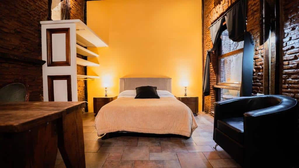 A imagem mostra o quarto do Pasaje Solar, um dos hotéis em San Telmo, Buenos Aires. O ambiente conta com uma cama grande ao centro, com mesa de cabeceira e luminárias de ambos os lados. De um lado há uma janela grande de vidro com cortinas e um poltrona preta, do outro, uma prateleira aberta e uma mesa, o chão é de madeira e atrás da cama a parede é amarela.