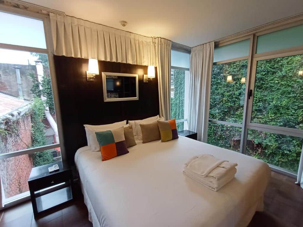 Vista do interior do quarto do Ribera Sur Hotel, em San Telmo, Buenos Aires, com cama de casal ao dentro, cabeceira alta com quadro e duas luminárias, há portas e janelas de vidros de ambos os lados em que é possível ver a vegetação do lado de fora. Há cortinas para garantir a privacidade.