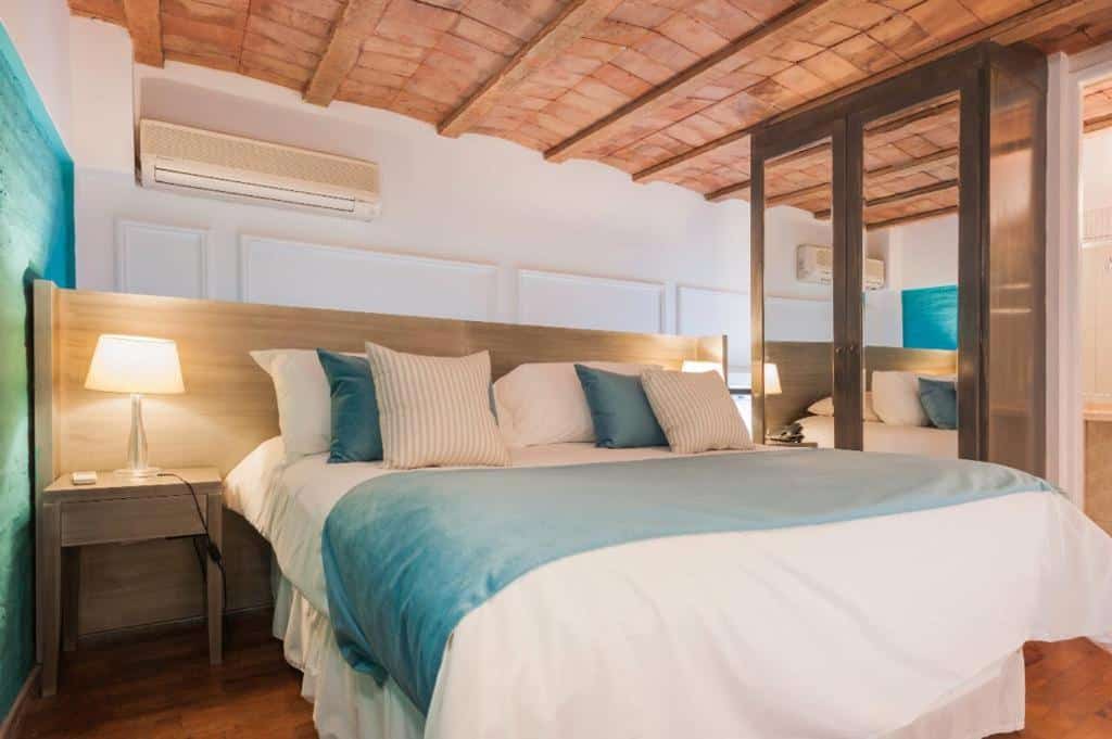 Quarto do San Telmo Suites com cama de casal grande no centro, com mesinha de cabeceira e luminária de ambos os lados. De um lado tem um guarda-roupa com espelho nas portas, o teto é de madeira e há um ar-condicionado.