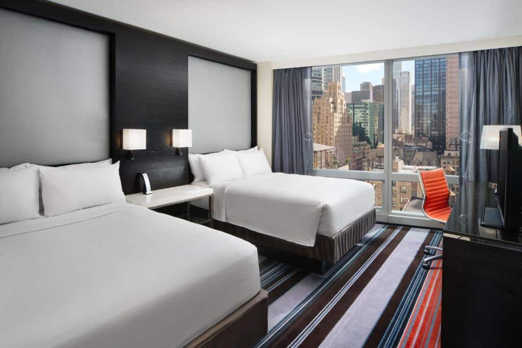 No quarto do Courtyard by Marriott New York Manhattan há duas camas de casal com roupas de cama brancas, lado a lado. À direita, há uma mesa de trabalho preta com uma cadeira de escritório laranja. A parede do fundo é uma grande janela com vista para os prédios de Nova York.