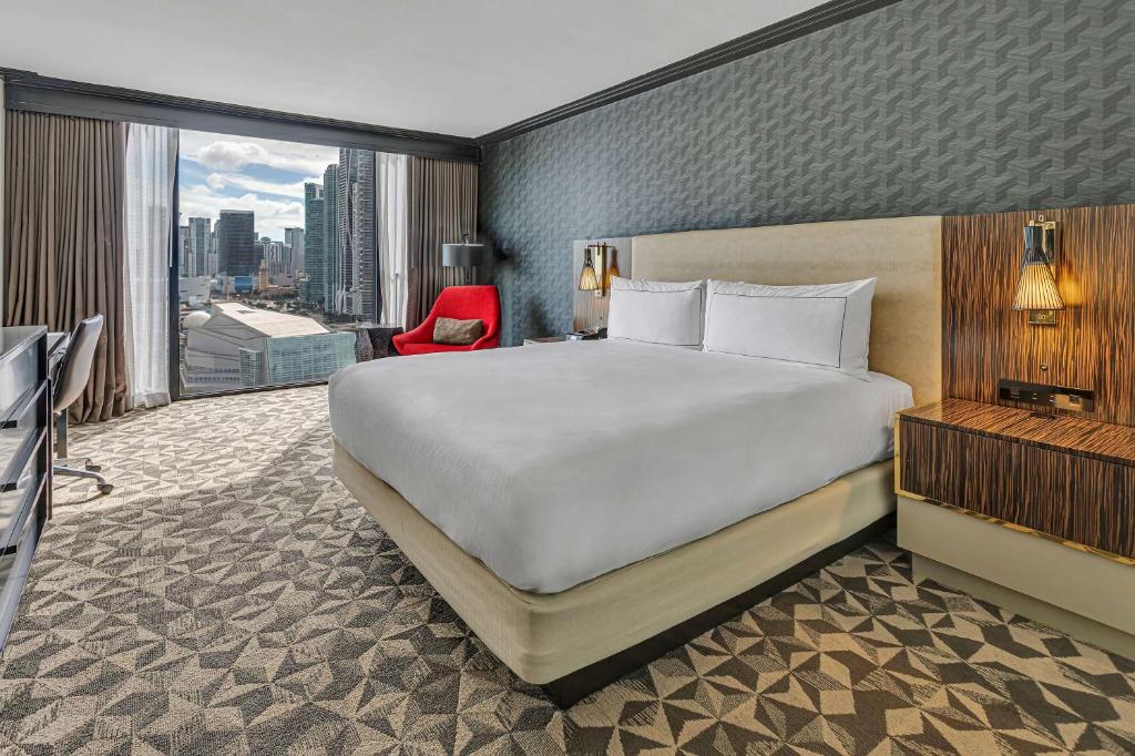Quarto do Hilton Miami Downtown com cama de casal no centro do quarto e em cada lado da cama uma cômoda do lado esquerdo uma poltrona e em frente a cama uma cômoda. . Representa hotéis no centro de Miami.