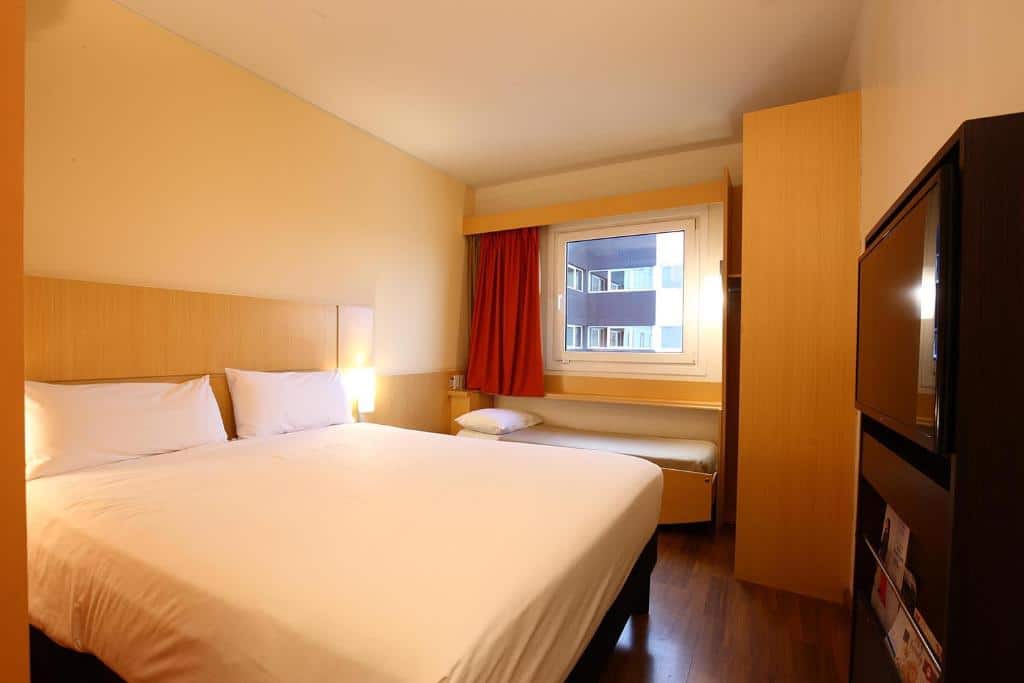O quarto do Ibis Buenos Aires Obelisco é compacto e funcional, com uma cama de casal centralizada, complementada por um design simples e moderno. As paredes possuem um tom suave, criando uma atmosfera acolhedora. Ao lado da cama, há uma janela que permite a entrada de luz natural, com cortinas em tom vermelho. O quarto também conta com uma televisão de tela plana montada na parede em frente à cama, e um pequeno espaço de trabalho com uma cadeira. O ambiente é prático, ideal para uma estadia confortável e sem excessos.