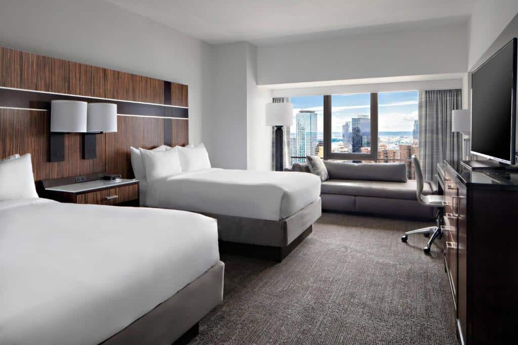 No quarto New York Marriott Marquis Hotel há duas camas de casal com cabeceiras de madeira escura. À direita, há um sofá cinza próximo à janela. Na parede ao fundo, há uma grande janela com vista para a cidade. No canto esquerdo, há uma mesa de trabalho com uma cadeira de escritório.