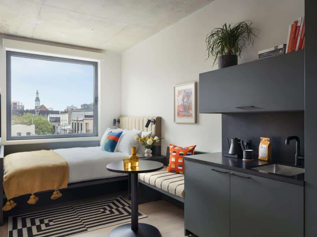 No quarto do Penny Williamsburg Hotel há uma cama de casal com colcha amarela e almofadas coloridas. À direita da cama, uma pequena mesa redonda preta com um vaso dourado em cima. Ao fundo, uma janela grande com vista para a cidade. À esquerda, há uma bancada preta com uma pia e uma cafeteira. Um dos hotéis onde ficar em Nova York.