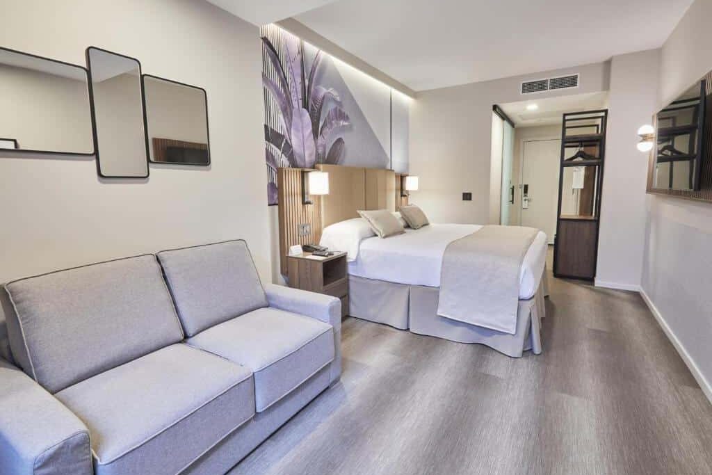 No quarto do Riu Plaza Manhattan Times Square há uma cama de casal ao centro, com cabeceira estofada. À esquerda da cama, há uma mesa de trabalho com um espelho acima e uma TV presa na parede. À direita, uma poltrona e uma mesa de cabeceira com um abajur. A janela ao fundo oferece vista para a cidade. Um dos hotéis onde ficar em Nova York.