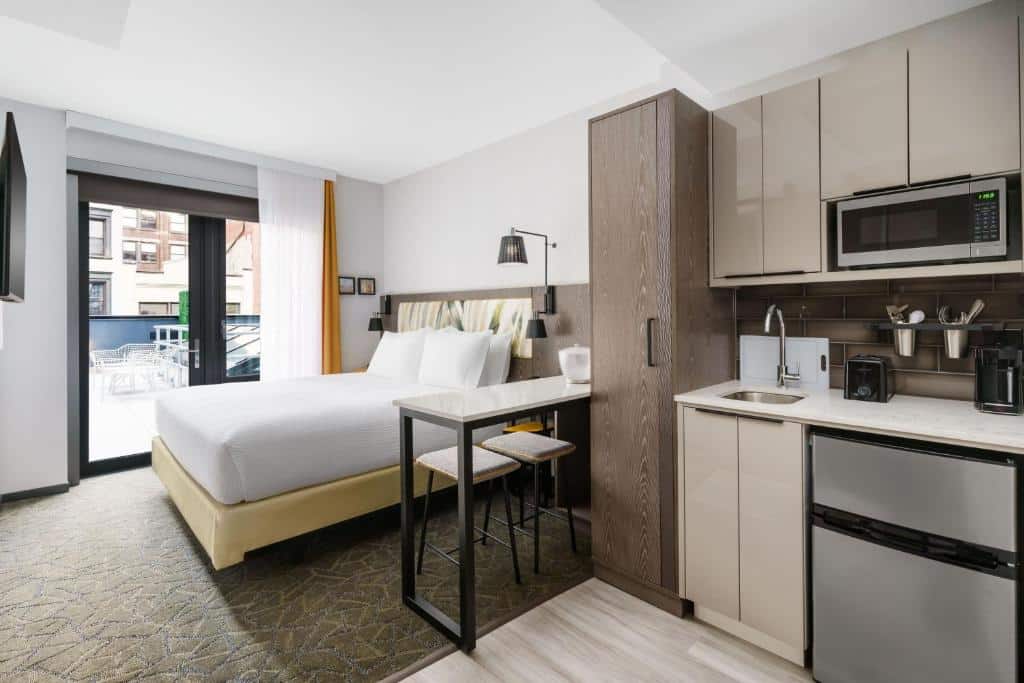 No quarto do TownePlace Suites by Marriott New York Manhattan Chelsea há uma cama de casal com cabeceira estofada, ao centro. À esquerda, uma pequena cozinha com armários, micro-ondas e pia. À direita, uma mesa de trabalho com uma cadeira preta. Na parede do fundo, uma grande janela com cortinas. Um dos hotéis onde ficar em Nova York.