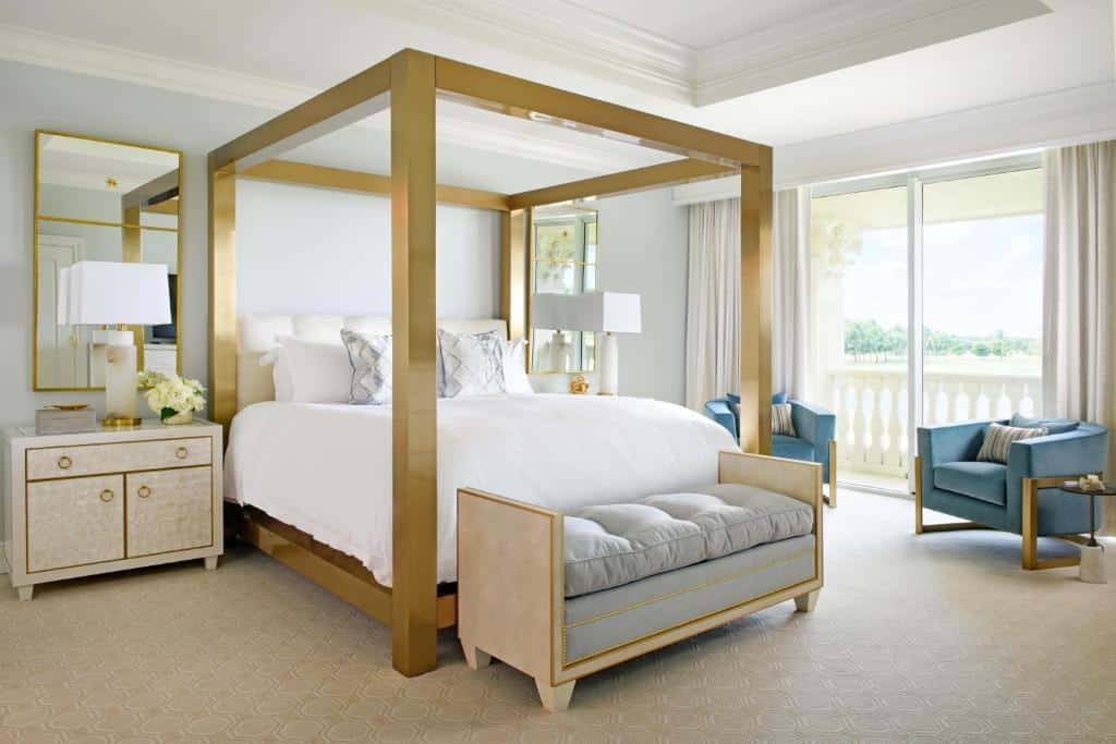 Quarto luxuoso do Trump National Doral Golf Resort com cama de dossel dourada, roupa de cama branca, duas cadeiras azuis, uma mesa lateral e uma grande janela com cortinas. Representa onde ficar em Miami.