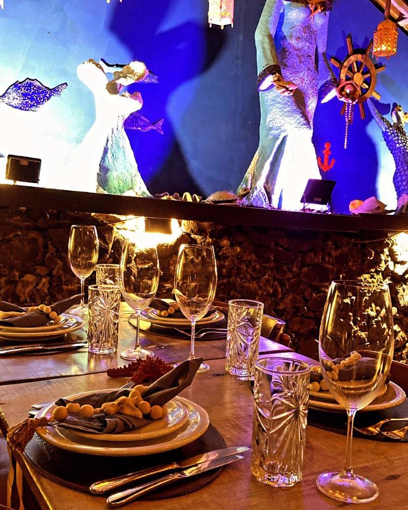 Foto do Cacimba Bistrô, em que mostra uma mesa de madeira com pratos, taças, talheres e copos. Há uma parede e pedra no fundo enfeitada com figuras regionais.