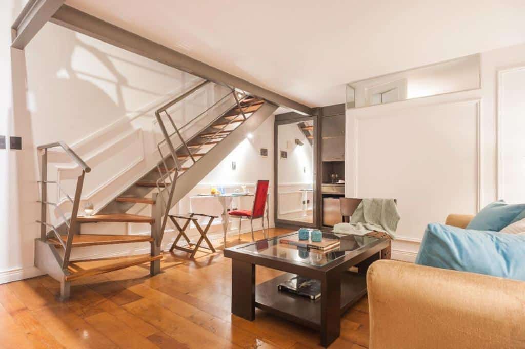 Sala do San Telmo Suites, em Buenos Aires, com sofá e almofadas azuis, mesinha de centro com tampo de vidro, uma escrivaninha atrás com cadeira, um espelho de corpo inteiro e um armário grande. Por fim, uma escada indica que há outro andar no ambiente.