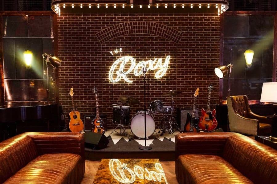 No palco do Roxy Hotel há uma bateria no centro, cercada por guitarras. À frente do palco, há dois sofás de couro marrom, um de cada lado de uma mesa de centro retangular. Na parede de fundo, há um letreiro iluminado com o nome "Roxy".