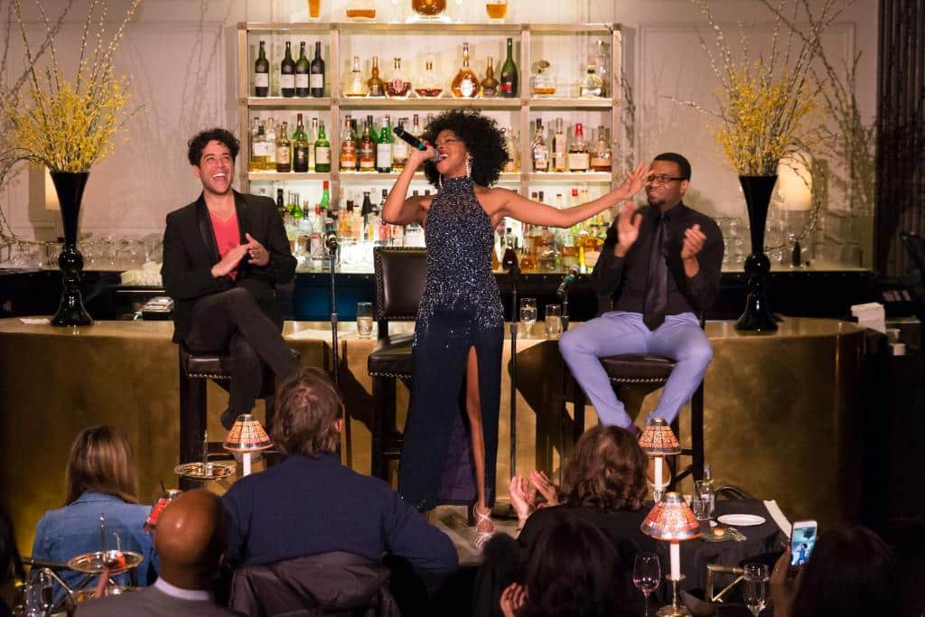 No palco do The Pierre Hotel há uma cantora ao centro, segurando um microfone. À esquerda e à direita dela, há duas pessoas sentadas em bancos altos, aplaudindo. Ao fundo, há uma estante com várias garrafas e enfeites. O público está sentado em mesas redondas em frente ao palco.