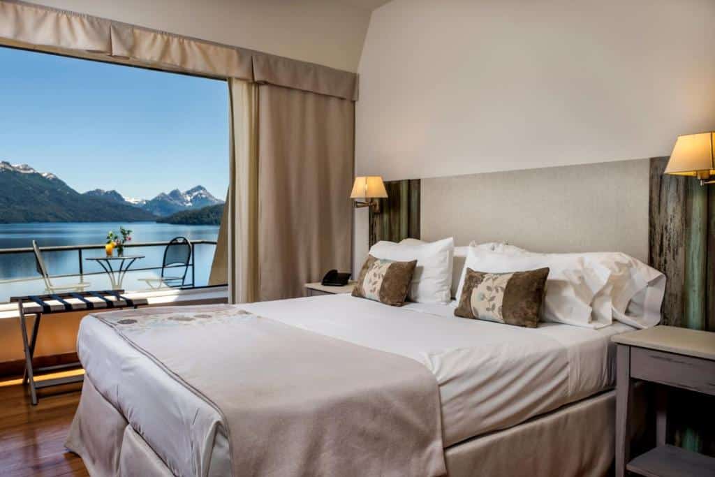 Quarto do Sol Arrayan Hotel & Spa. Uma cama de casal do lado direito com uma estante e um abajur de cada lado. No fundo uma parede de vidro com vista da varanda e do lago.