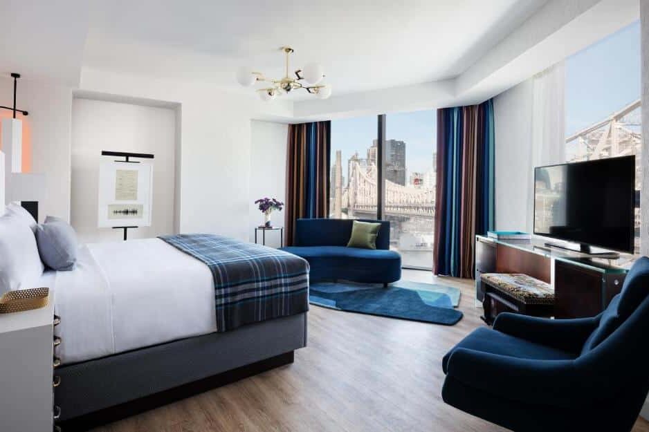 Na suíte do Graduate New York Hotel há uma cama de casal ao centro, com cabeceira estofada e roupa de cama branca. À direita, uma poltrona azul e uma TV sobre um suporte. À esquerda, uma pequena mesa de cabeceira com um abajur. A janela ao fundo oferece uma vista da cidade.