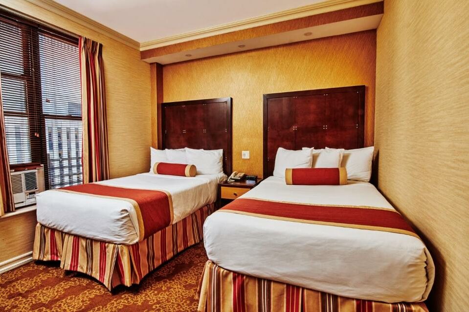 No quarto do Radio City Apartments há duas camas de solteiro com cabeceiras de madeira escura, uma ao lado da outra, com um corredor estreito entre elas. Cada cama tem uma almofada cilíndrica vermelha. À esquerda, uma janela com persianas. O ambiente é decorado com papel de parede bege e detalhes em vermelho e dourado nas roupas de cama e cortinas.