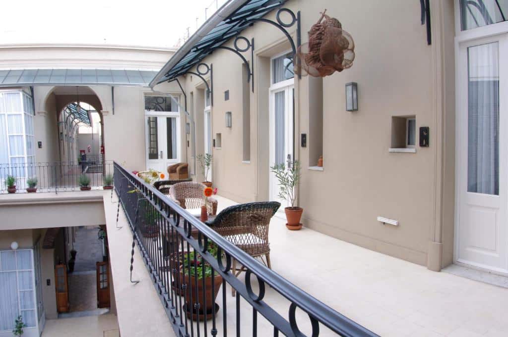 A imagem mostra uma varanda do hotel Patios de San Telmo, com um design clássico e elegante. A varanda é coberta e decorada com móveis de vime, incluindo cadeiras confortáveis e uma mesa pequena, criando um espaço acolhedor para relaxar. Há plantas em vasos que adicionam um toque de verde ao ambiente, e a varanda oferece uma vista interna para outras partes do edifício, que é caracterizado por sua arquitetura tradicional com detalhes em ferro forjado nas grades e estruturas de suporte. A área é iluminada, transmitindo uma sensação de tranquilidade e charme histórico.