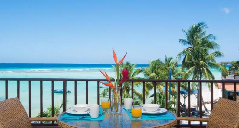 Varanda do Yellow Bird Hotel com uma mesa posta na varanda com uma flor. No fundo a vista do mar com palmeiras, para representar hotéis em Barbados.