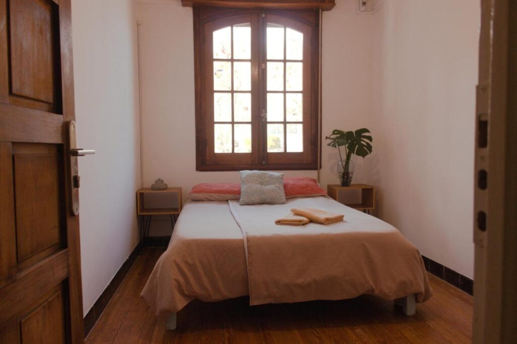 Quarto do Youki Haus Hostel, com uma cama no centro e uma janela em cima, de cada lado uma estante de apoio.