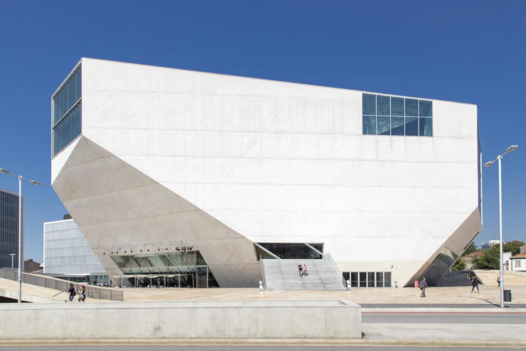 Imagem da Casa da Música em um edifício geométrico branco moderno no Porto, com grandes janelas e uma entrada pública, situado em frente a um céu azul claro.