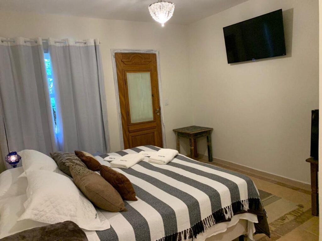 Quarto do Aldeia Parque Pousada Rural. Uma cama de casal no canto esquerdo, de frente uma TV. No undo uma porta e uma janela com cortina.