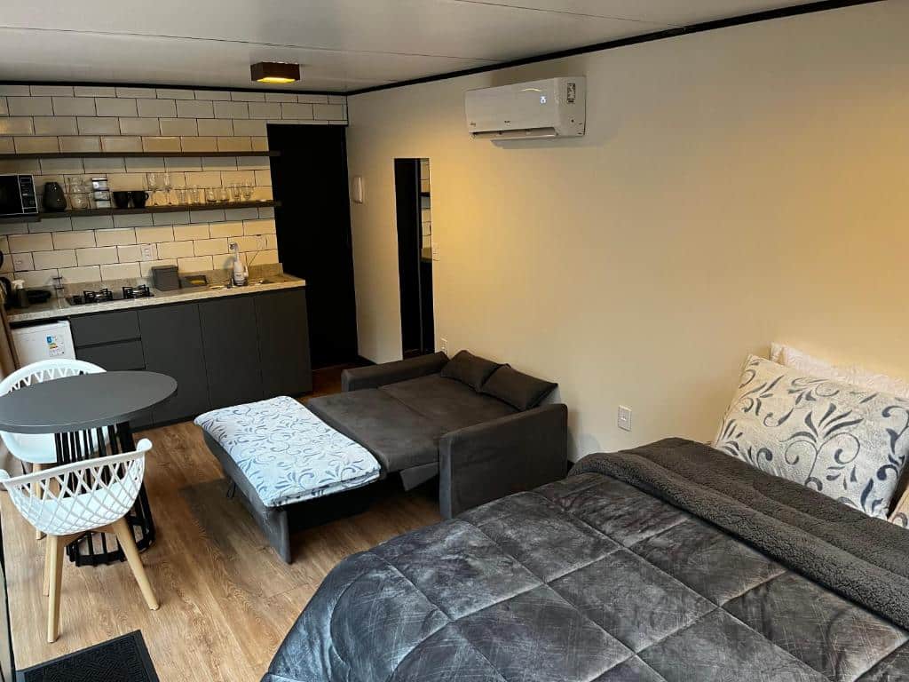 O quarto na imagem é um estúdio moderno que combina áreas de estar e dormir. À direita, há uma cama com um edredom cinza escuro e almofadas estampadas. À esquerda da cama, há um sofá seccional em tecido cinza claro, posicionado de frente para o centro do quarto. Em frente ao sofá, há um puff com tecido semelhante ao do sofá.
Uma pequena mesa de jantar redonda com duas cadeiras brancas está situada perto da área da cozinha. A kitchenette parece estar totalmente equipada, com armários superiores e inferiores em branco, bancadas escuras e eletrodomésticos de aço inoxidável, incluindo um micro-ondas acima do que parece ser um forno ou lava-louças.