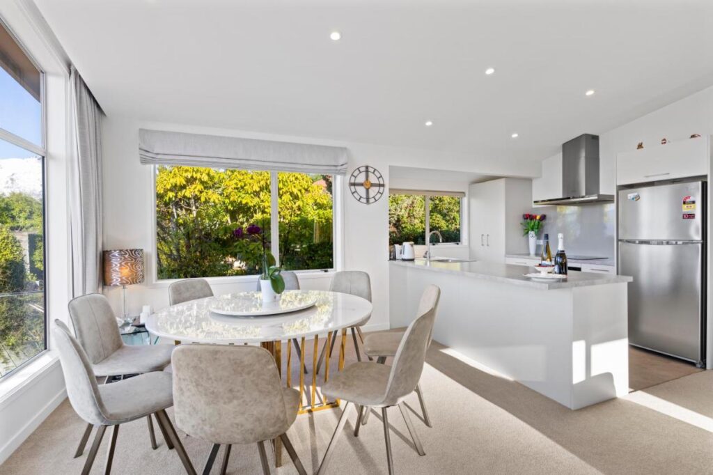 Cozinha moderna do Alpine Home, aluguel de temporada em Queenstown. Uma área de jantar com mesa redonda, cadeiras, janelas grandes, armários brancos e uma geladeira de aço inoxidável compõe o ambiente.