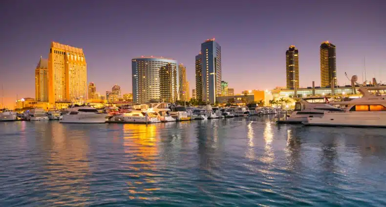 Foto da baía de San Diego para o post de como chegar em San Diego. Em primeiro plano está a água e, ao fundo, vários tipos de barcos e prédios iluminados, já que a foto foi tirada à noite.