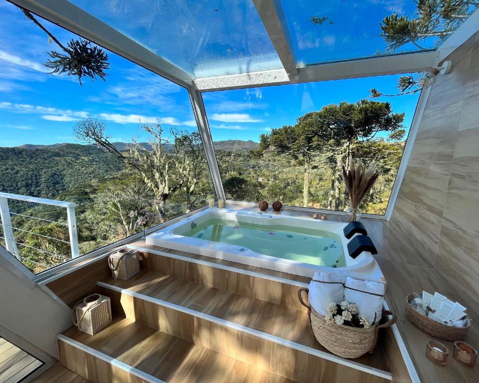 Banheira de hidromassagem no Benta Rosa Chalés de luxo em Urubici, com deck de madeira em um ambiente todo revestido de vidro, dos lados e acima, mantendo o valor interior enquanto é possível observar a natureza ao redor, com céu azul, nuvens e muitas araucárias.