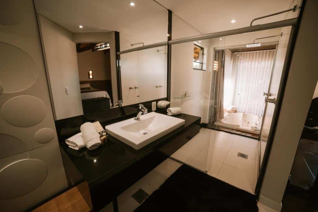 Banheiro amplo do Virá Charme Resort, com pia com espaço abaixo, espelho grande, box também espaçoso com chuveiro e barra de apoio, que conecta ainda com uma banheira de hidromassagem mais ao fundo.