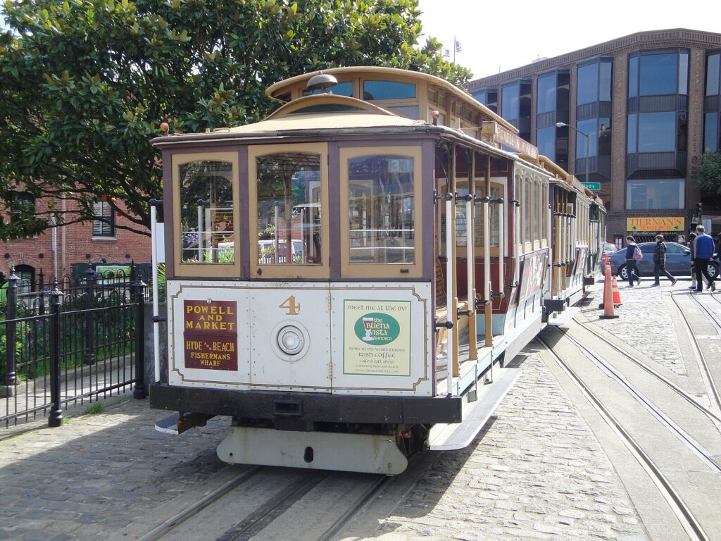 Um bonde clássico de São Francisco parado nos trilhos, com um prédio de tijolos e árvores ao fundo.