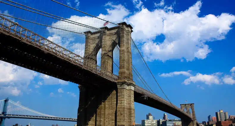 A imagem pra ilustrar o post sobre o que fazer no Brooklyn mostra a Ponte do Brooklyn, uma famosa ponte suspensa em Nova York. Ela atravessa o Rio East, conectando os bairros de Manhattan e Brooklyn. A foto destaca uma das torres de pedra da ponte, com seus arcos góticos característicos e uma rede de cabos de aço que sustentam o tabuleiro da ponte. O céu está claro, com poucas nuvens, sugerindo que é um dia ensolarado.