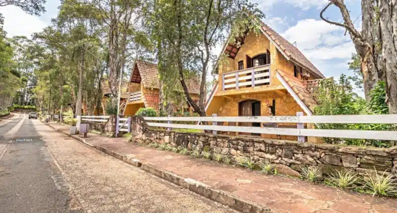 A imagem mostra chalés em estilo alpino, construídos com pedras de tons amarelos e telhados inclinados de telhas vermelhas. Árvores altas e verdes cercam a área. Trata-se do Miraguá Refúgios Pousada, para representar chalés para casal no interior de São Paulo.