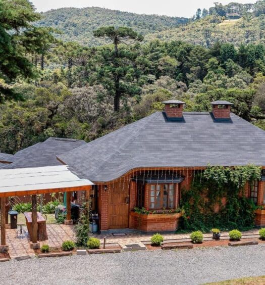 A imagem mostra uma pousada rústica rodeada por uma paisagem montanhosa e muita vegetação. A construção principal tem paredes de tijolos vermelhos, telhado escuro com chaminés e janelas com molduras de madeira, decoradas com flores. Na parte frontal, há plantas trepadeiras subindo pela parede, e um pátio de tijolos que leva à entrada. Ao lado da pousada, uma área coberta oferece um espaço ao ar livre com mesa e cadeiras. Ao fundo, árvores altas e colinas completam a vista, criando um ambiente tranquilo e natural. O local faz parte da lista de chalés na Serra da Mantiqueira.