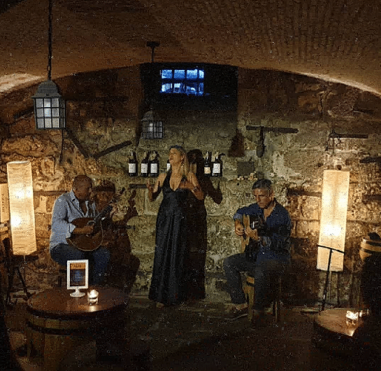 Imagem da Casa do Fado com três músicos estão se apresentando em um ambiente rústico e com pouca iluminação, com paredes de pedra, garrafas de vinho e iluminação suave.