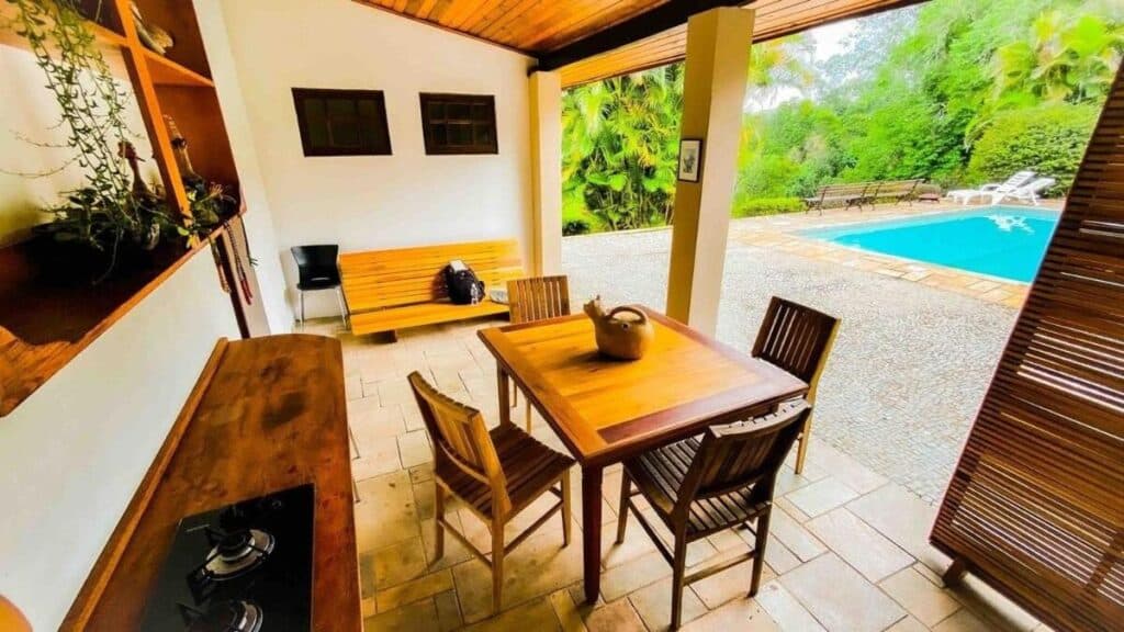 Área externa do Chalé com vista incrível, um dos chalés em São Roque. Uma mesa com quatro cadeiras no centro, do lado esquerdo uma bancada com cooktop. Do lado direito, no fundo, um piscina.