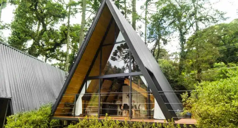 A imagem mostra um chalé em formato triangular com uma grande fachada de vidro, cercado por árvores altas e vegetação densa. A estrutura tem um design moderno, com moldura em preto e interior visível, com tons de madeira clara que dão um ar acolhedor. Há uma varanda de madeira com guarda-corpo de metal, e cortinas brancas que decoram o ambiente. O chalé está localizado em uma área arborizada, dando a impressão de estar imerso na natureza, proporcionando um refúgio tranquilo e privativo. Este é o Chalé Mon Amour, uma das recomendações de chalés em Campos do Jordão.