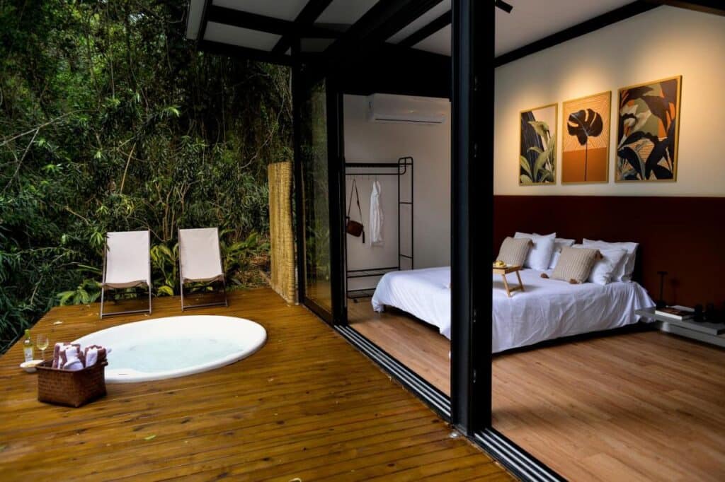 Do lado direito o quarto do Chalé na mata 201, do lado esquerdo, na varanda com porta de vidro, uma banheira e duas cadeiras.