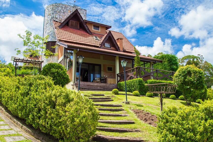 A imagem mostra uma casa de campo grande e charmosa, o Chalé dos Sonhos, com telhado inclinado de telhas vermelhas e uma estrutura de pedra em uma das extremidades. O ambiente é cercado por um jardim bem cuidado, com arbustos verdes e uma escadaria de pedra que leva até a entrada principal. A casa tem janelas amplas e uma varanda de madeira, com uma bela vista para o jardim. O céu ao fundo está azul com algumas nuvens brancas, e o cenário passa uma sensação de tranquilidade e natureza. Esse local é uma recomendação entre os chalés em Campos do Jordão.