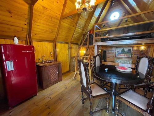 A imagem mostra o interior de um chalé de madeira com decoração rústica nos Chalés Caiçara. No centro, há uma mesa redonda de madeira escura com quatro cadeiras de estilo clássico. À esquerda, há uma geladeira retrô vermelha ao lado de uma pequena pia de madeira. O teto alto de madeira inclinado segue o formato triangular do chalé, com um mezanino acessado por uma escada de madeira ao fundo. Uma luminária pendente ilumina o ambiente, que é acolhedor e tem um toque vintage. Na parede, há um quadro decorativo com paisagem.